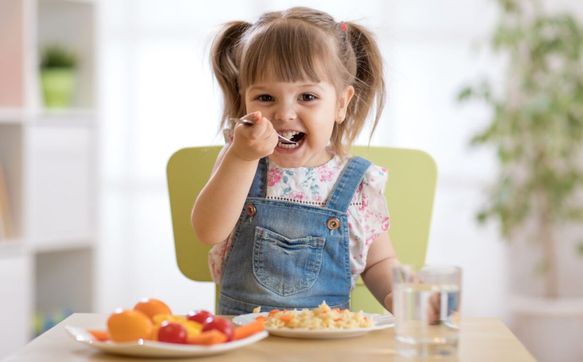 Photo shows a little girl eating a spoon of plant-based dinner, primarily fruit and vegetables, with a glass of milk. A new study finds that vegan children can experience several health benefits from the diet, but that fortified foods - such as soy milk - are key