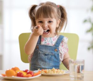 Photo shows a little girl eating a spoon of plant-based dinner, primarily fruit and vegetables, with a glass of milk. A new study finds that vegan children can experience several health benefits from the diet, but that fortified foods - such as soy milk - are key