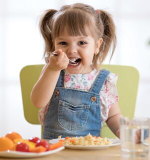 Photo shows a little girl eating a spoon of plant-based dinner, primarily fruit and vegetables, with a glass of milk. A new study finds that vegan children can experience several health benefits from the diet, but that fortified foods - such as soy milk - are key