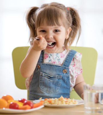 Photo shows a little girl eating a spoon of plant-based dinner, primarily fruit and vegetables, with a glass of milk. A new study finds that vegan children can experience several health benefits from the diet, but that fortified foods - such as soy milk - are key