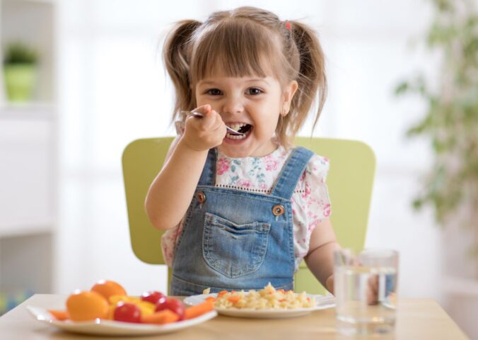 Photo shows a little girl eating a spoon of plant-based dinner, primarily fruit and vegetables, with a glass of milk. A new study finds that vegan children can experience several health benefits from the diet, but that fortified foods - such as soy milk - are key