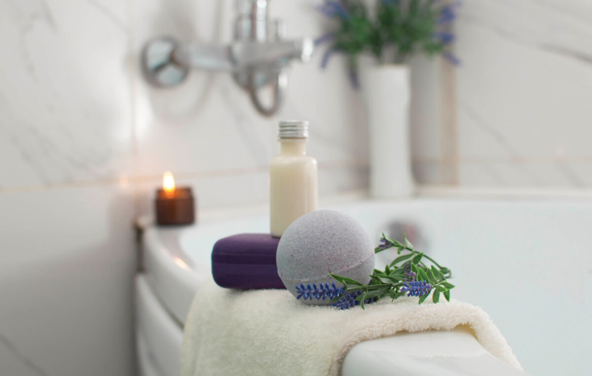 Photo shows a bath bomb-type product alongside soap, lavender, and a candle on the edge of a bath