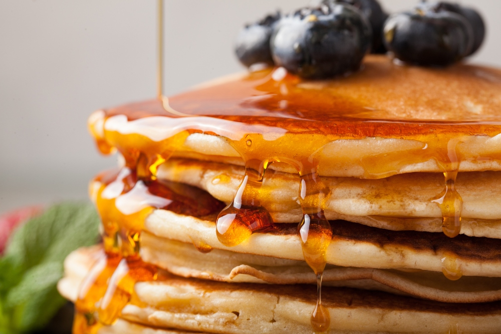 Close-up of pancakes with fresh blueberries, dripping maple syrup, on a light background, to illustrate article about the healthiest vegan sweeteners.