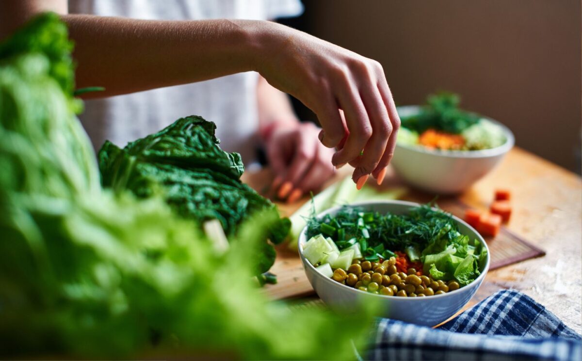 Hand pours green onions in a bowl with green peas, cucumbers, carrots, lettuce and dill standing on a table, to illustrate article about higher NAD+ levels in vegans