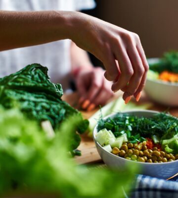 Hand pours green onions in a bowl with green peas, cucumbers, carrots, lettuce and dill standing on a table, to illustrate article about higher NAD+ levels in vegans