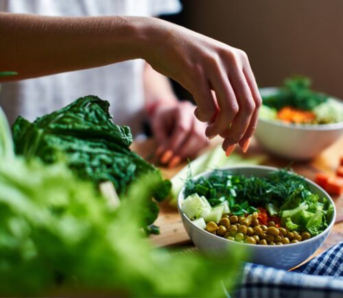 Hand pours green onions in a bowl with green peas, cucumbers, carrots, lettuce and dill standing on a table, to illustrate article about higher NAD+ levels in vegans