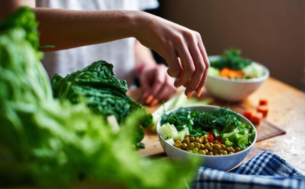 Hand pours green onions in a bowl with green peas, cucumbers, carrots, lettuce and dill standing on a table, to illustrate article about higher NAD+ levels in vegans