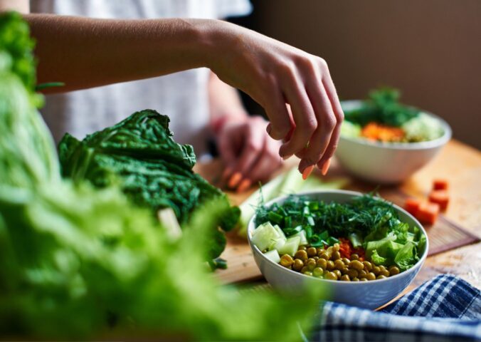 Hand pours green onions in a bowl with green peas, cucumbers, carrots, lettuce and dill standing on a table, to illustrate article about higher NAD+ levels in vegans