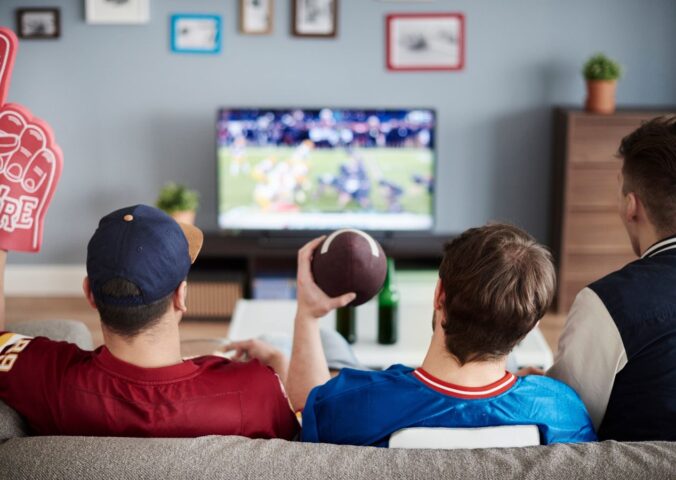 Photo shows three men on a sofa facing away from the camera and watching American football. According to a new survey, many Americans will be eating chicken wings during this Sunday's Super Bowl, and most of them would consider a plant-based or vegan alternative