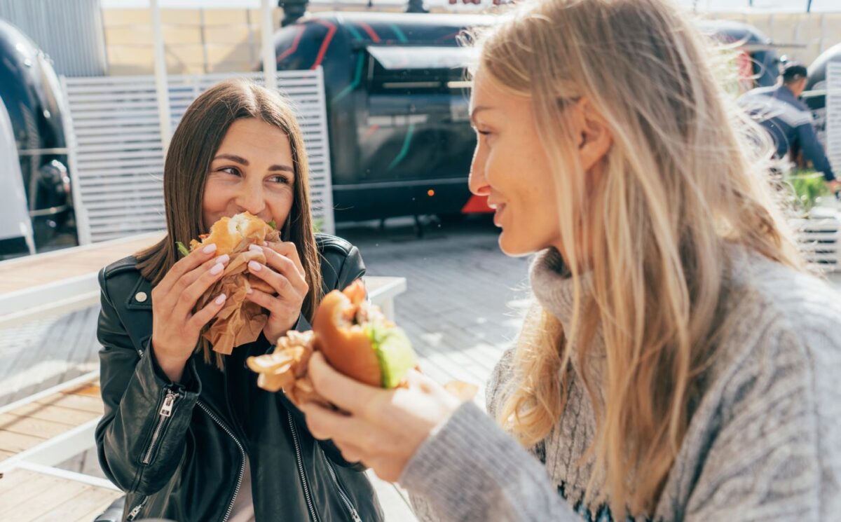 Photo shows two women laughing and eating lunch in an outdoor restaurant area with foodtrucks. London was recently named the best vegan city in the UK and in the world