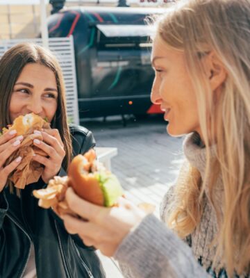 Photo shows two women laughing and eating lunch in an outdoor restaurant area with foodtrucks. London was recently named the best vegan city in the UK and in the world