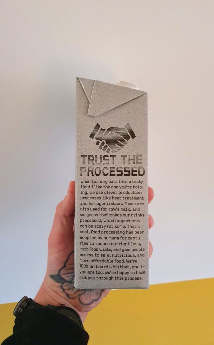 Photo shows someone's hand as they hold up a carton of Oatly with the new "trust the processed" label addressing the oat milk's classification as a processed food
