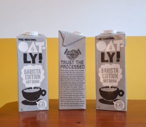 Photo shows someone's hand as they hold up a carton of Oatly with the new "trust the processed" label addressing the oat milk's classification as a processed food