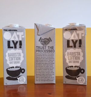 Photo shows someone's hand as they hold up a carton of Oatly with the new "trust the processed" label addressing the oat milk's classification as a processed food