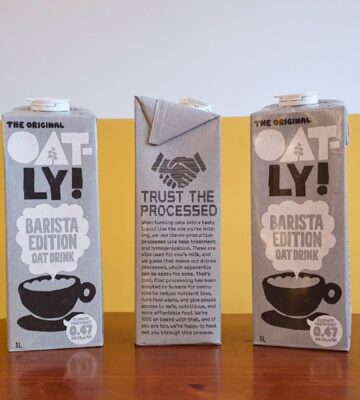 Photo shows someone's hand as they hold up a carton of Oatly with the new "trust the processed" label addressing the oat milk's classification as a processed food