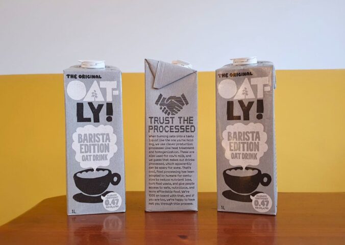 Photo shows someone's hand as they hold up a carton of Oatly with the new "trust the processed" label addressing the oat milk's classification as a processed food