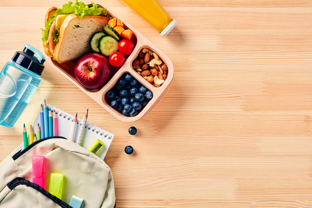 Photo shows a compartmentalized lunch box filled with fruit, nuts, salad, and a sandwich next to a school bag, coloring pencils, and a water bottle
