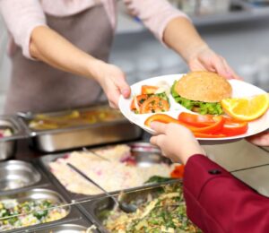 Photo shows a cafeteria worker handing a school child a tray of vegetarian food, including a burger and vegetables. A US Senator has just sponsored the Plant-Powered School Meals Pilot Act.