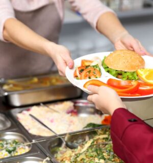 Photo shows a cafeteria worker handing a school child a tray of vegetarian food, including a burger and vegetables. A US Senator has just sponsored the Plant-Powered School Meals Pilot Act.