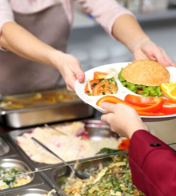 Photo shows a cafeteria worker handing a school child a tray of vegetarian food, including a burger and vegetables. A US Senator has just sponsored the Plant-Powered School Meals Pilot Act.