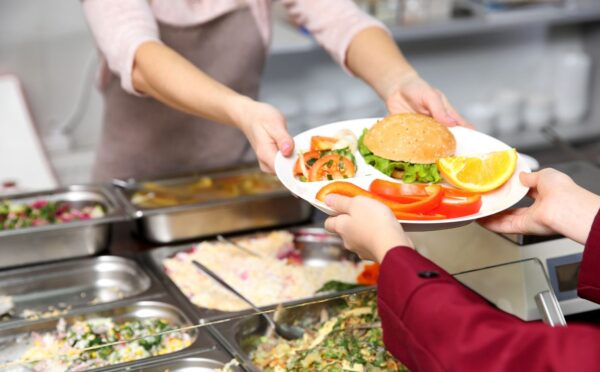 Photo shows a cafeteria worker handing a school child a tray of vegetarian food, including a burger and vegetables. A US Senator has just sponsored the Plant-Powered School Meals Pilot Act.