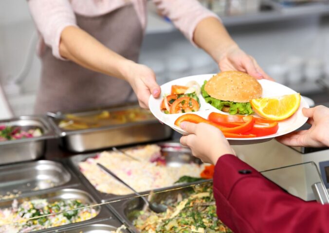 Photo shows a cafeteria worker handing a school child a tray of vegetarian food, including a burger and vegetables. A US Senator has just sponsored the Plant-Powered School Meals Pilot Act.