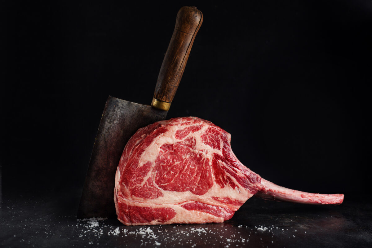 Photo shows a tomahawk steak next to a cleaver on a black surface