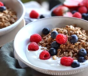 Bowls of homemade granola and plant-based yogurt topped with blueberries and raspberries, with maple syrup on the side, to illustrate article about budget-friendly plant-based recipes