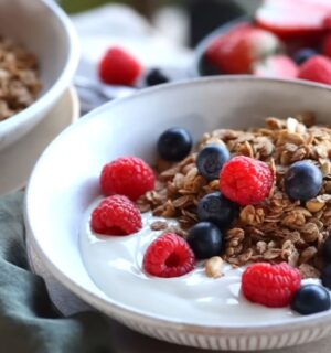 Bowls of homemade granola and plant-based yogurt topped with blueberries and raspberries, with maple syrup on the side, to illustrate article about budget-friendly plant-based recipes