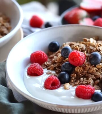 Bowls of homemade granola and plant-based yogurt topped with blueberries and raspberries, with maple syrup on the side, to illustrate article about budget-friendly plant-based recipes