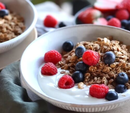 Bowls of homemade granola and plant-based yogurt topped with blueberries and raspberries, with maple syrup on the side, to illustrate article about budget-friendly plant-based recipes