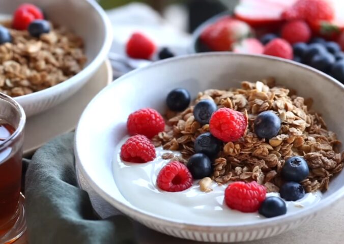 Bowls of homemade granola and plant-based yogurt topped with blueberries and raspberries, with maple syrup on the side, to illustrate article about budget-friendly plant-based recipes