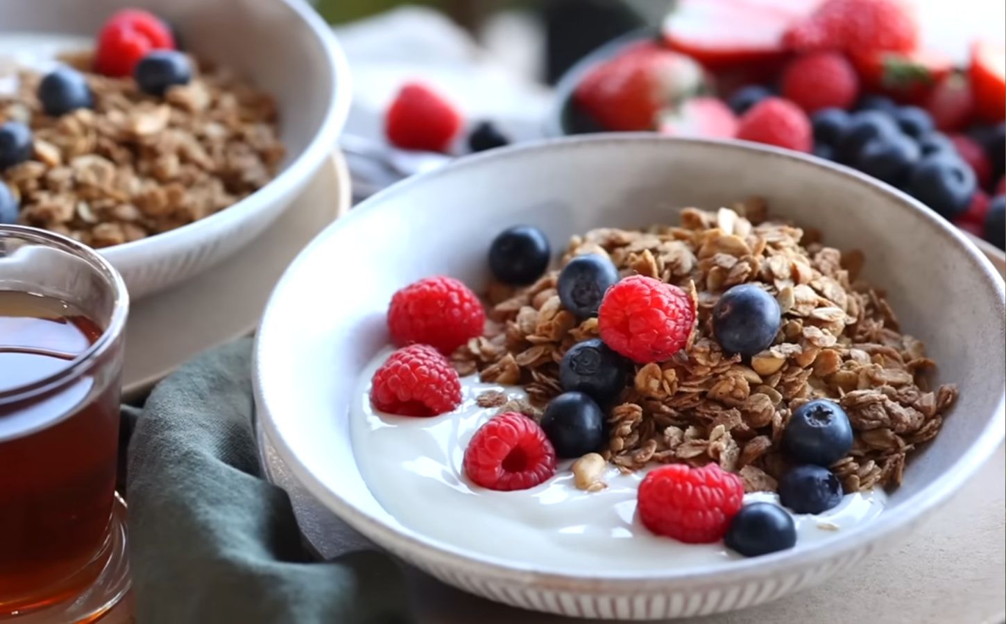 Bowls of homemade granola and plant-based yogurt topped with blueberries and raspberries, with maple syrup on the side, to illustrate article about budget-friendly plant-based recipes