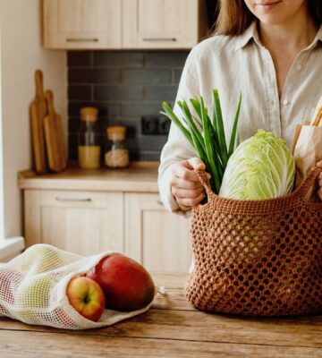 Woman unpacking groceries on her kitchen counter, with one bag full of veg and a baguette, and another of fruit, to illustrate article about how meal timing can benefit health