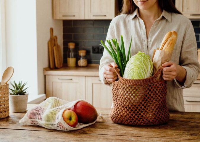 Woman unpacking groceries on her kitchen counter, with one bag full of veg and a baguette, and another of fruit, to illustrate article about how meal timing can benefit health