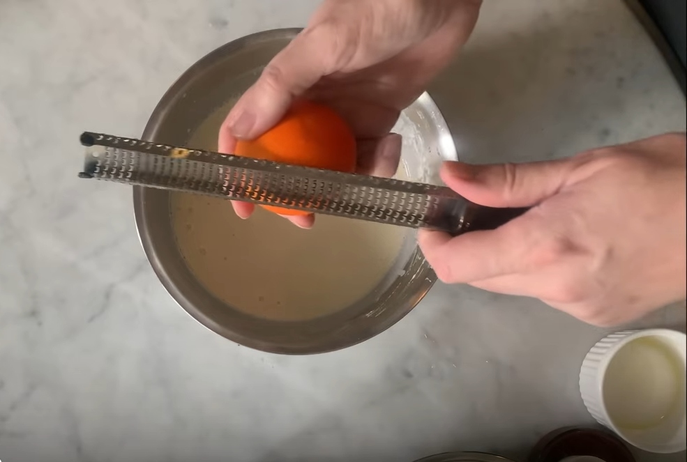 Close-up of Roudaut’s hands grating fresh organic orange zest into a mixing bowl, with ingredients for his vegan orange zest crepes visible in the background