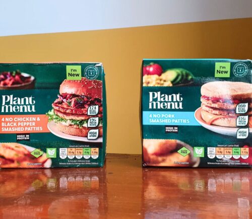 Photo shows the two "smashed" burger products from Aldi - the No Pork Smashed Patties and the No Chicken & Black Pepper Smashed Patties - on a wooden dining table against a yellow and white background