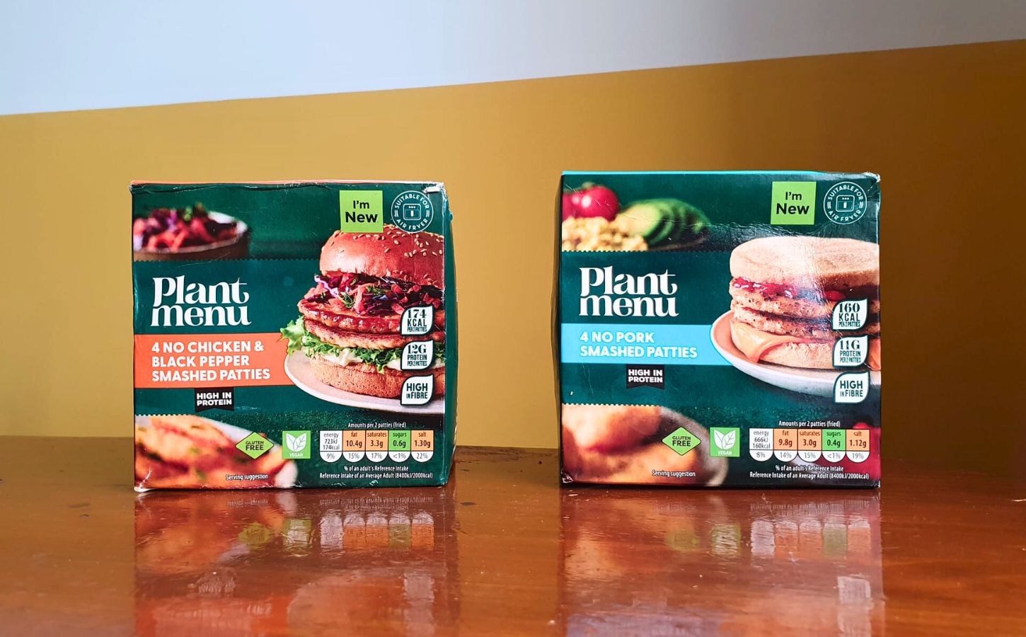 Photo shows the two "smashed" burger products from Aldi - the No Pork Smashed Patties and the No Chicken & Black Pepper Smashed Patties - on a wooden dining table against a yellow and white background