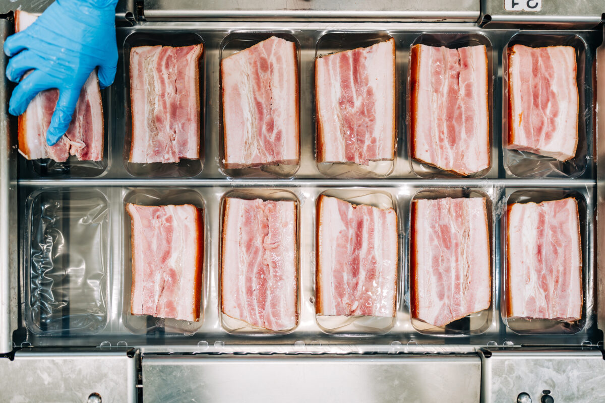 Photo shows a factory worker in blue gloves arranging pieces of pork before they are cut into thing bacon slices