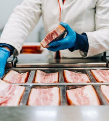 Photo shows a factory worker in blue gloves arranging pieces of pork before they are cut into thing bacon slices. How is bacon made?