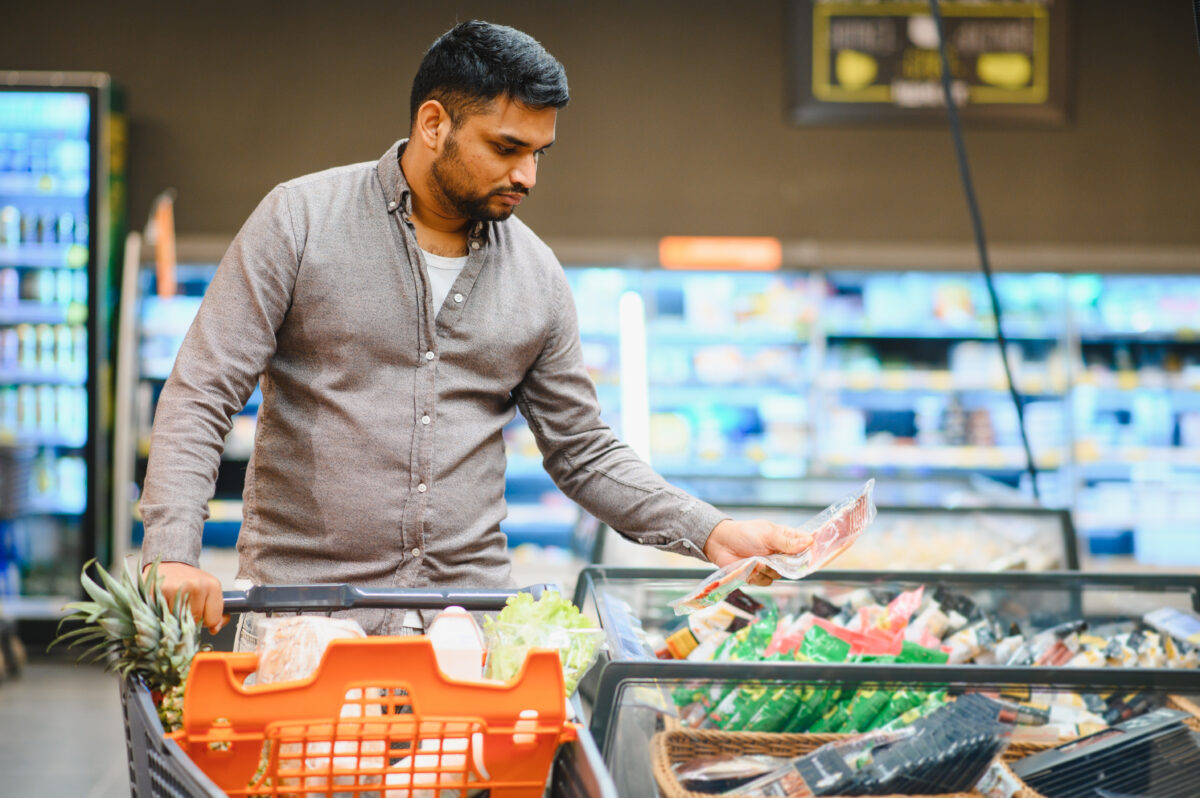 Photo shows a young man shopping in a supermarket and holding up a packet of meat. 