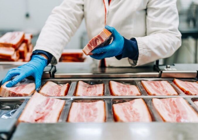 Photo shows a factory worker in blue gloves arranging pieces of pork before they are cut into thing bacon slices. How is bacon made?