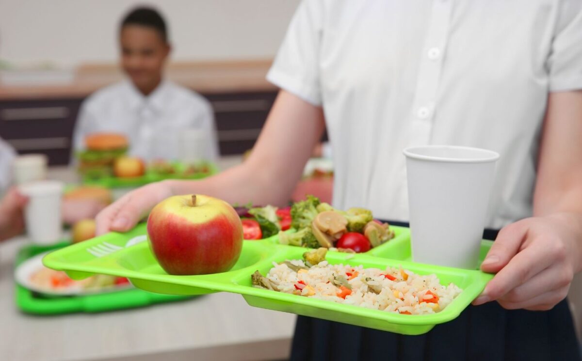 Photo shows a school child in uniform holding a green plastic tray with plant-based food on it, including an apple, grains, fresh fruit, and salad. Children and young people in Poland are now guaranteed access to plant-based school meals for the first time ever