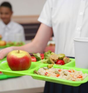 Photo shows a school child in uniform holding a green plastic tray with plant-based food on it, including an apple, grains, fresh fruit, and salad. Children and young people in Poland are now guaranteed access to plant-based school meals for the first time ever