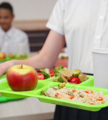 Photo shows a school child in uniform holding a green plastic tray with plant-based food on it, including an apple, grains, fresh fruit, and salad. Children and young people in Poland are now guaranteed access to plant-based school meals for the first time ever