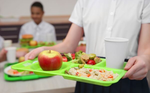 Photo shows a school child in uniform holding a green plastic tray with plant-based food on it, including an apple, grains, fresh fruit, and salad. Children and young people in Poland are now guaranteed access to plant-based school meals for the first time ever