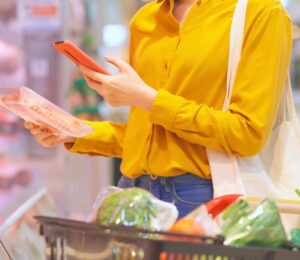 Photo shows a young woman holding a pack of processed meat in one hand and looking at her phone in the other while shopping in a supermarket. Sales of nitrite-cured bacon have dropped significantly as consumers look to avoid the controversial additive over its links to cancer risk.