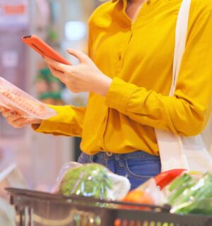 Photo shows a young woman holding a pack of processed meat in one hand and looking at her phone in the other while shopping in a supermarket. Sales of nitrite-cured bacon have dropped significantly as consumers look to avoid the controversial additive over its links to cancer risk.