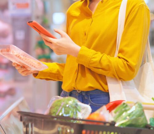 Photo shows a young woman holding a pack of processed meat in one hand and looking at her phone in the other while shopping in a supermarket. Sales of nitrite-cured bacon have dropped significantly as consumers look to avoid the controversial additive over its links to cancer risk.