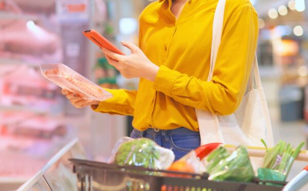 Photo shows a young woman holding a pack of processed meat in one hand and looking at her phone in the other while shopping in a supermarket. Sales of nitrite-cured bacon have dropped significantly as consumers look to avoid the controversial additive over its links to cancer risk.